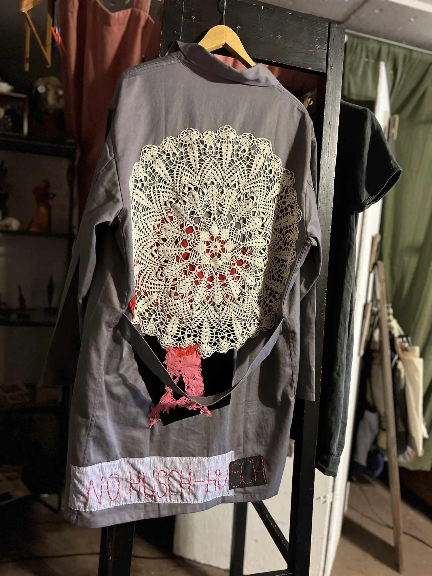 Grey coat with a large crocheted lace doily across the back, NO HUSCH-HUSCH hand-lettered along the hem